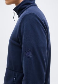 Πλάγια όψη ατόμου που φοράει μπλε ναυτικό φλις μπουφάν με ψηλό γιακά και φερμουάρ, δείχνοντας το λογότυπο του μαμούθ στο πάνω μέρος του μανικιού.