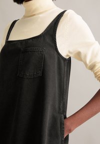 Zwarte denim jurk met een vierkante halslijn, zijzakken en een borstzak, gedragen over een lichtgekleurde turtleneck. Gladde textuur.