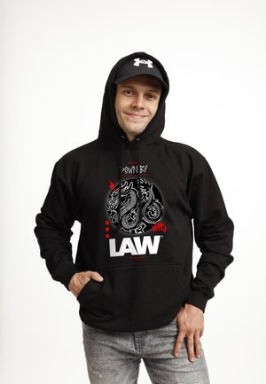 Młody mężczyzna w czarnej bluzie z kapturem z grafiką smoka i napisem „LAW”, czarną czapką i szarymi dżinsami, pozujący z ręką w kieszeni.