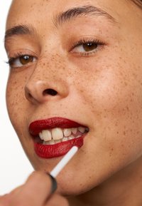 Röd flytande läppstift appliceras med en applikator på modellens läppar, som visar upp fräknig hud, vita tänder och en slät, matt finish.