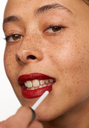 Un rouge à lèvres liquide rouge est appliqué avec un applicateur sur les lèvres d'un modèle, mettant en valeur une peau tachetée de rousseur, des dents blanches et un fini lisse et mat.