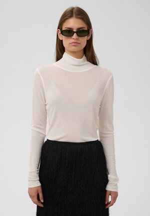 Femme portant des lunettes de soleil noires étroites, un haut à col roulé blanc à manches longues, et une jupe noire texturée, debout devant un fond clair uni.