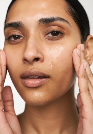 Primo piano di un volto con pelle idratata, caratterizzata da una texture liscia, una leggera lucentezza, un incarnato marrone chiaro e sopracciglia scure. Le mani toccano delicatamente il viso.