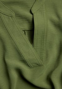 Olivgrüner Stoff mit V-Ausschnitt, der zwei schräg verlaufende Blenden und dezente Nähdetails aufweist, glatte Textur und keine sichtbaren Muster.