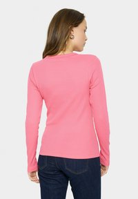 Roze ribgebreide longsleeve shirt met een ronde halslijn, getailleerd ontwerp en zachte textuur. Gemaakt om te dragen met donkerblauwe jeans, waarbij een gladde achterkant zichtbaar is.