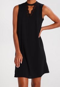 Robe noire sans manches avec un décolleté en V et des détails en dentelle. Coupe évasée, texture lisse, longueur genoux et finition légèrement brillante.