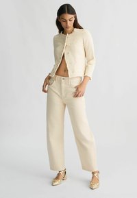 Veste en denim beige clair et courte avec des bords effilochés, boutons argentés, associée à un pantalon taille haute assorti. Les chaussures sont des ballerines dorées.
