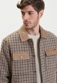 Bruine en crèmekleurige geruite jas met een corduroy kraag en zakken, met drukknoopsluitingen en een gestructureerd, geweven stofdesign.