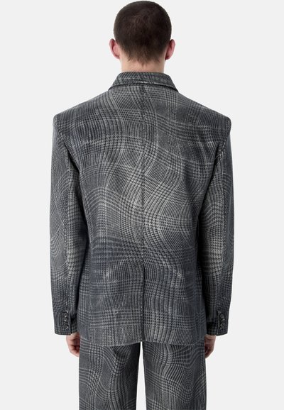 Hombre vistiendo un traje de cuadros en gris y negro, con chaqueta y pantalones a juego, visto desde atrás sobre un fondo blanco liso.