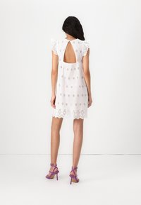 Witte jurk van knielengte met kanten afwerking, open rugontwerp en bloemmotieven. Model draagt paarse sandalen met bandjes.