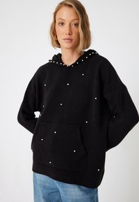 Schwarze Kapuzenjacke aus Strickstoff, mit einer vorderen Tasche und verstreuten weißen Perlenakzenten. Enthält eine Kapuze mit Zugband und Perlendetails.