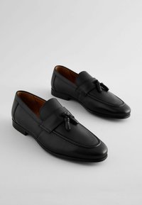Mocasines de cuero negro con acentos texturizados, que presentan un borlas decorativa. Superficie lisa y forma clásica, con un pequeño tacón.
