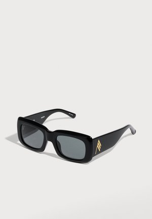 Schwarze rechteckige Sonnenbrille mit dunklen Gläsern und einem goldenen geometrischen Logo an den breiten Bügeln vor hellem Hintergrund.