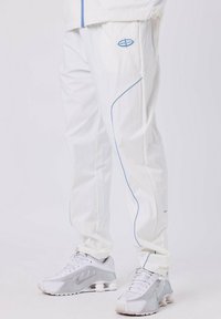 Personne portant un pantalon de survêtement blanc avec des liserés bleus et des chaussures de sport blanches avec des accents gris, se tenant sur un sol blanc.