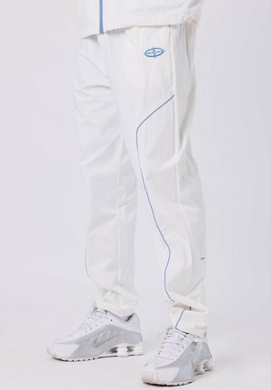Persona vestida con pantalones de chándal blancos con ribetes azules y zapatillas deportivas blancas con detalles grises, de pie sobre un suelo blanco.