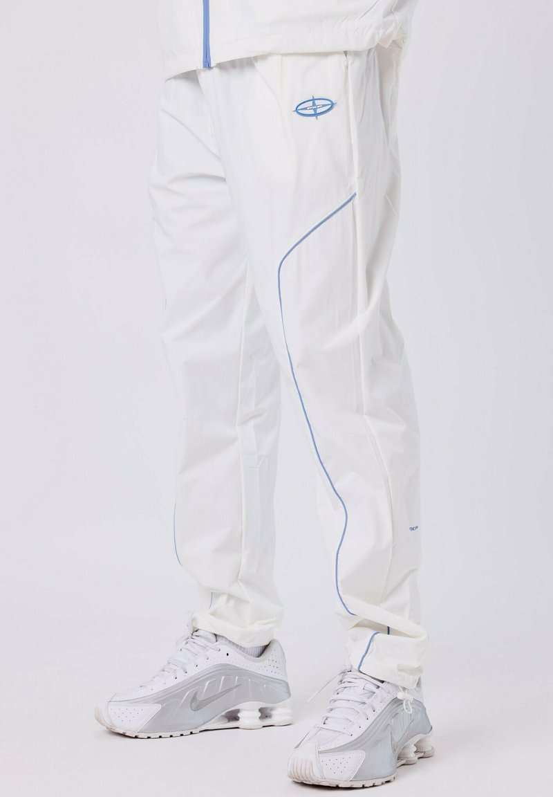 Personne portant un pantalon de survêtement blanc avec des liserés bleus et des chaussures de sport blanches avec des accents gris, se tenant sur un sol blanc.