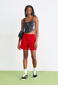 Top cropped preto com riscas brancas, calções vermelhos e sapatilhas pretas com atacadores creme. A modelo leva uma pequena mala de ombro preta.