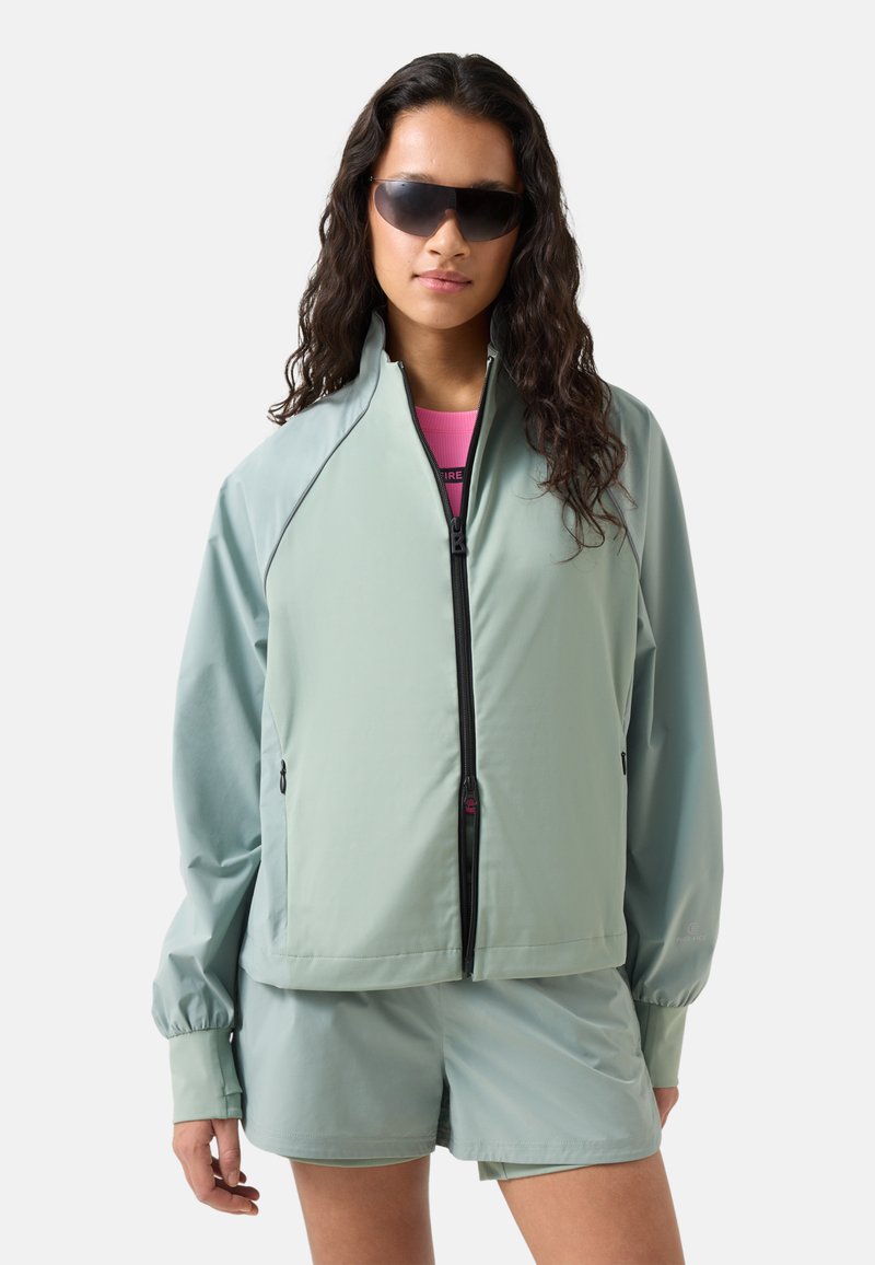 Kvinde iført stor lysegrøn lynlåstrøje og matchende shorts, med langt krøllet hår og store mørke solbriller.