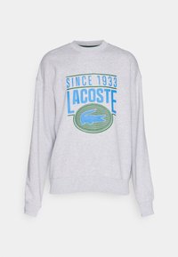 Felpa grigia chiara con colletto tondo, caratterizzata da una grafica verde e blu che riporta la scritta "Dal 1933 Lacoste" e un emblema di coccodrillo blu.