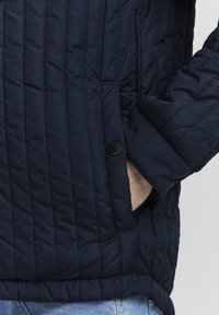 Giacca trapuntata blu navy con tasca laterale dotata di un bottone nero, polsini a costine e un tessuto liscio e testurizzato.