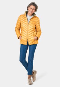 Veste matelassée jaune avec fermeture éclair, col montant et deux poches latérales. Associée à un jean bleu et des baskets beige clair.
