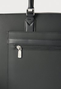 Sac cabas noir texturé avec poignée en cuir, poche avant zippée et tirette de fermeture éclair argentée arborant un petit logo crocodile en relief.