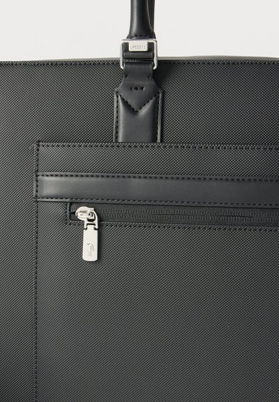 Sac cabas noir texturé avec poignée en cuir, poche avant zippée et tirette de fermeture éclair argentée arborant un petit logo crocodile en relief.