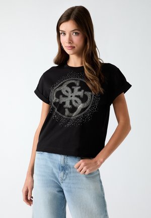 Giovane donna con lunghi capelli castani indossa una maglietta nera a maniche corte con polsini arrotolati e un logo grafico argentato, abbinata a jeans azzurri chiari.
