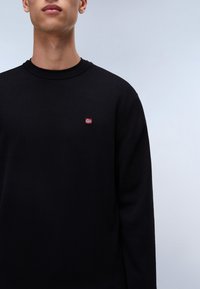 Zwarte crewneck trui van textiel met een kleine geweven Noorse vlag emblem op de borst. Eenvoudig design, lange mouwen.
