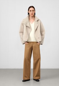 Beżowy płaszcz z owczej wełny z szerokim kołnierzem, nałożony na kremowy sweter z dekoltem w serek. Brązowe spodnie o szerokich nogawkach i czarne botki za kostkę dopełniają stylizację.