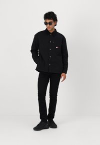 Cămașă neagră cu nasturi, guler și buzunar frontal, asortată cu pantaloni negri slim-fit și adidași negri, purtată de un model cu ochelari de soare.