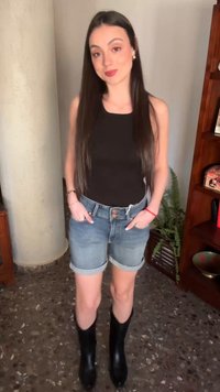 Νεαρή γυναίκα με μακριά σκούρα μαλλιά, ντυμένη με μαύρο αμάνικο τοπ, τζιν σορτς και μαύρες μπότες, στέκεται μέσα σε εσωτερικό χώρο με τα χέρια στις τσέπες.