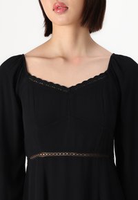 Melnā kleita ar kvadrātveida izgriezumu, garām pūkainām piedurknēm un mežģīņu apdari. Taisns griezums ar mīkstu tekstūru un smalkiem akcentiem jostasvietā.