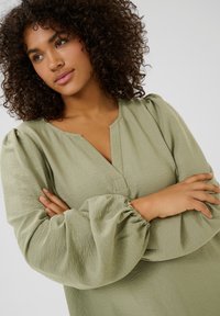 Blouse à manches longues, vert clair, avec un col en V et des manches bouffantes froncées. Fabriquée en tissu texturé. Présente une coupe décontractée.
