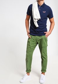 Tmavě modré polo tričko, zelené kapsáčové kalhoty s bočními kapsami a bílé tenisky. Model drží světle šedý svetr přehozený přes rameno.