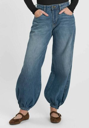 Donna che indossa pantaloni di jeans blu larghi con polsini elastici e scarpe basse marroni, mani nelle tasche, su uno sfondo semplice.