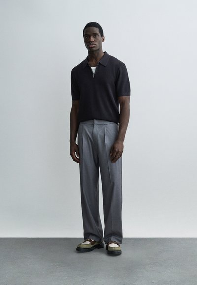 Άντρας που στέκεται φορώντας μαύρο πουκάμισο με κοντά μανίκια και γιακά, γκρι πλισέ παντελόνι και μπεζ δετά παπούτσια, σε απλό φόντο.