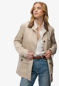 Mujer ajustándose una chaqueta beige con cuello marrón sobre una camisa blanca, con cinturón trenzado marrón y jeans azules, mirando hacia su izquierda.
