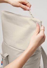 Hands with rings unzipping a beige fabric backpack with a front pocket and strap against a plain background.