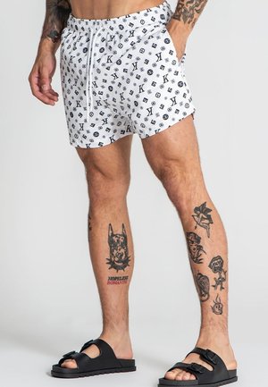 Mužské nohy s tetovaniami, oblečené v bielych šortkách s vzorom a čiernych šľapkách na jednoduchej svetlej ploche.