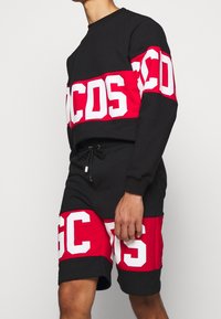 Homme portant un sweat-shirt noir et rouge et un short assorti, tous deux imprimés de grandes lettres blanches en blocs.