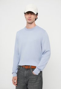 Μακρυμάνικο πουλόβερ γαλάζιου χρώματος με ριμπ μανσέτες; γκρι τζιν; καφέ ζώνη; λευκό καπέλο με κεντημένο το «BOSS». Απλή, καθημερινή εμφάνιση.