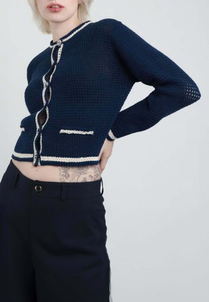 Persona che indossa un cardigan lavorato a uncinetto blu scuro con bordo bianco e pantaloni neri, mano appoggiata sulla parte bassa della schiena, tatuaggio visibile sulla pelle esposta.