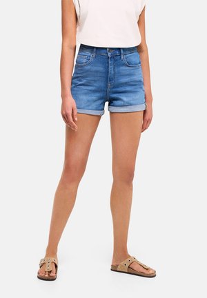 Mujer con pantalones cortos vaqueros azules con dobladillo enrollado, una camiseta blanca sin mangas y sandalias beige sin cordones, de pie frente a un fondo sencillo.
