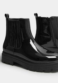 Stivali alla caviglia in pelle verniciata nera con dettagli rigati, cerniere laterali e suole in gomma spessa, caratterizzati da una finitura liscia e lucida.