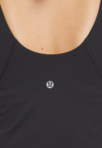 Gros plan d'un top de sport noir avec un col rond et un logo blanc Lululemon centré sous le col sur peau nue.