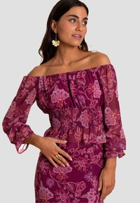 Paarse bloemen top met een off-the-shoulder ontwerp, smock taille, volant mouwen en een doorschijnende textuur. Gouden ring accent op de hand.