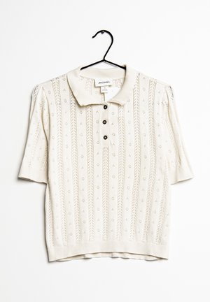 Kremowa dzianinowa koszulka polo z krótkim rękawem, z pionowymi koronkowymi wzorami, ściągaczowym dołem i mankietami, trzema czarnymi guzikami, zawieszona na czarnym wieszaku.