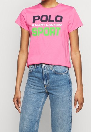 Kobieta w jaskraworóżowej koszulce z napisem "POLO RALPH LAUREN SPORT" w kolorze czarnym, białym i zielonym, w połączeniu z jeansami z wysokim stanem w niebieskim kolorze.