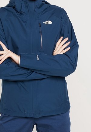 Marineblauwe, waterdichte North Face-jas met kap, gedragen door een persoon met gekruiste armen, waarbij de rits op de borst en het Gore-Tex-logo op de mouw zichtbaar zijn.